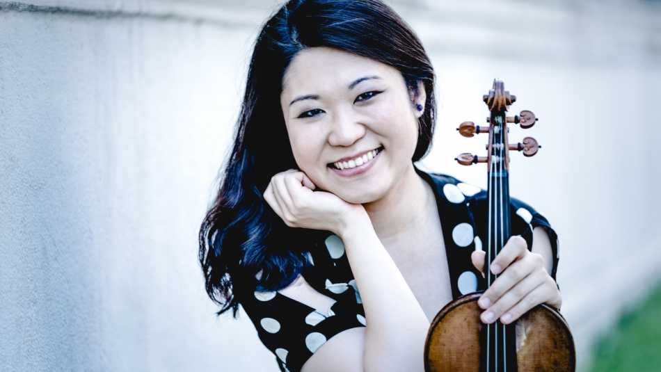 Tianwa Yang trägt ein gepunktetes Oberteil. Sie hält ihre Geige in der linken Hand, den Kopf auf die rechte gestützt und lächelt in die Kamera