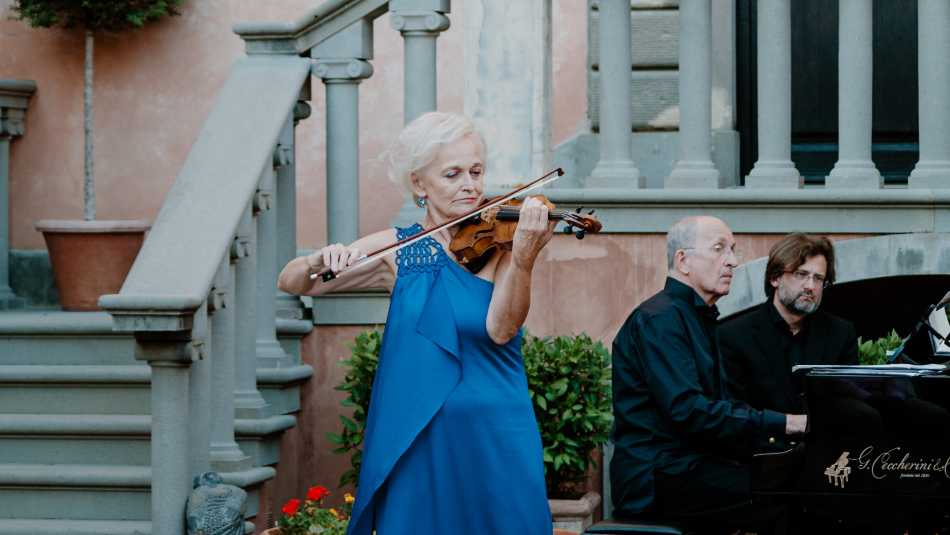 Annemarie Ortner spielt im blauen Kleid Geige. Im Hintergrund eine Treppe und ihr Mann, der sie am Klavier begleitet. 
