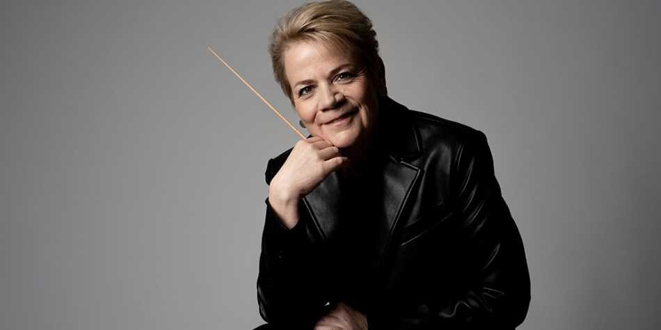 Marin Alsop in einer schwarzen Lederjacke, hält ihren Dirigentinnenstab in der rechten Hand, auf die sie auch ihr Kinn gestützt hat